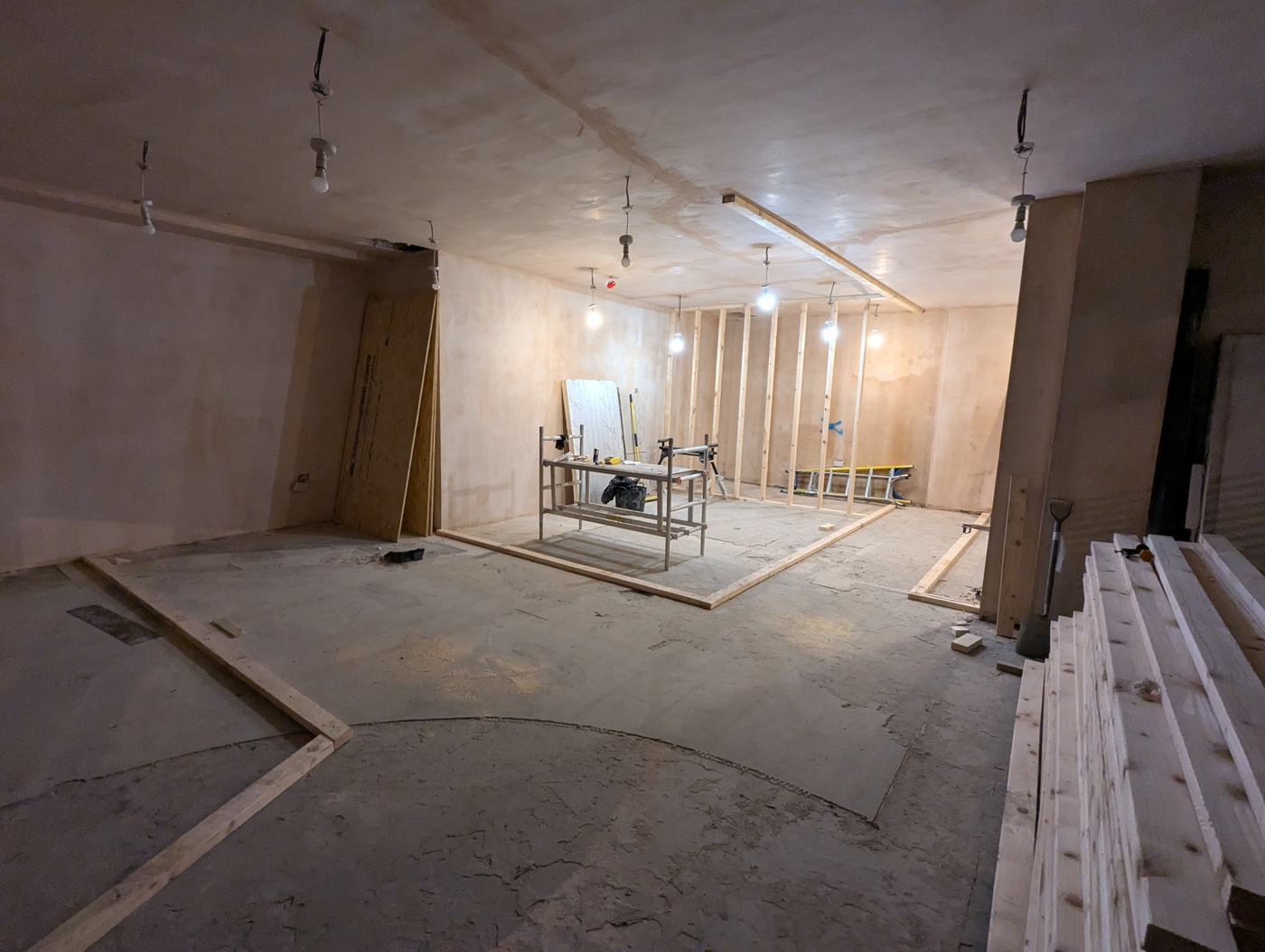 An empty room with a bare concrete floor, timber sole plates laid out marking where new stud walls will go, and a scaffold tower in the middle